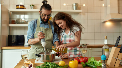 Italian man, chef cook using hand blender while preparing a meal. Young woman, girlfriend in apron pouring olive oil in the pot, helping him in the kitchen