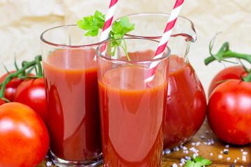 Fresh red tomato juice in a glass with a straw and jar with tomatoes on light wooden background.