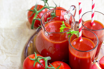 Fresh red tomato juice in a glass with a straw and jar with tomatoes on light wooden background.