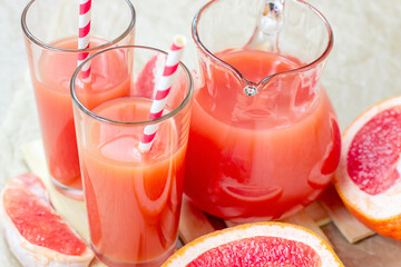 Fresh red grapefruit juice in a glass with a straw and jar with fruit pieces on light wooden background.