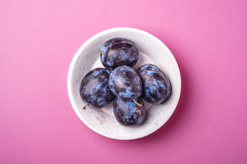 Fresh ripe plum fruits in white wooden bowl on pink minimal background, top view