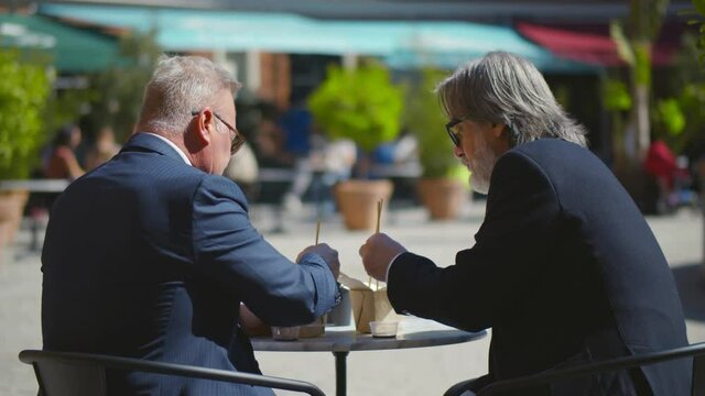 Back view of two successful elder executives sitting in outdoors cafe eating takeaway street food