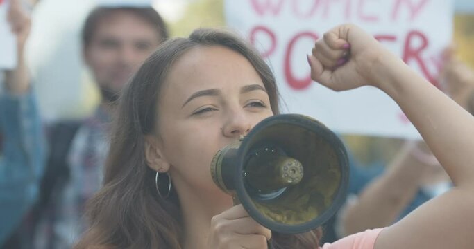 Close-up of young Caucasian feminist shouting through megaphone. Protest leader encouraging protesters with anti-sexism placards at the background. People protesting inequality. Cinema 4k ProRes HQ.