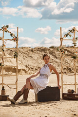 a young girl in a white lace boho chic dress poses on a sandy beach sitting on a vintage suitcase