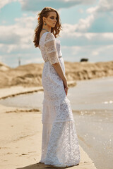 Fototapeta na wymiar A beautiful model poses on a deserted beach in a white lace dress on a Sunny day