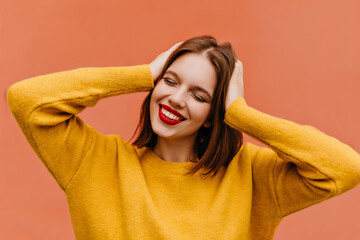 Wonderful european model expressing happiness in studio. Inspired caucasian girl in soft yellow sweater smiling with eyes closed.