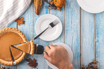 Overhead flat lay shot of a man's hand using a pie server to lift a freshly cut slice of pumpkin pie out of the pie tin with empty plates on a rustic blue painted wooden table surface.