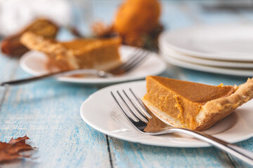 Closeup shot of a slice of pumpkin pie on a plate with a fork with plates and other objects blurred in the background on a blue painted rustic wooden table surface
