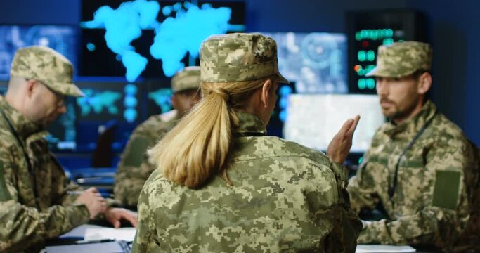 Rear of female general giving orders to mixed-races males militarian council at table. Deciding war or defense tactics in monitoring room. Back view Woman and multiethnic officers discussing terrorism