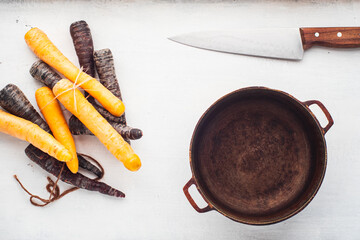 Purple and Yellow Carrots next to a Steel Pot and a Knife on a White Table