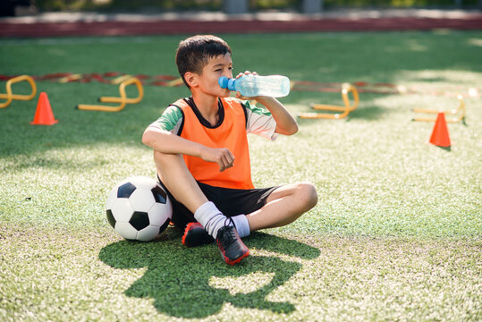 Tired teen boy in soccer uniform drinks with water from plastic bottle after intensive training at stadium in morning. - Powered by Adobe