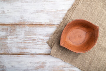 Empty clay brown bowl on white wooden background and linen textile. Top view, copy space.
