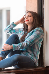 
White-skinned Latina woman with long hair, smiling while sitting on the edge of the window, wearing a plaid shirt and jeans