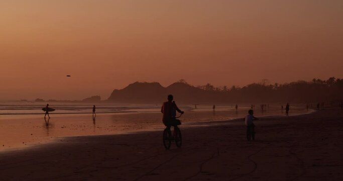 Father cycles by with his sone while on vacation along beach as golden sun sets in the distance 