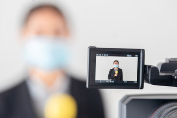 Fototapeta premium Young asian journalist woman wearing medical mask talking with microphone reporting news of COVID-19 pandemic and tracking the virus situation on camera screen. Focus on camera 