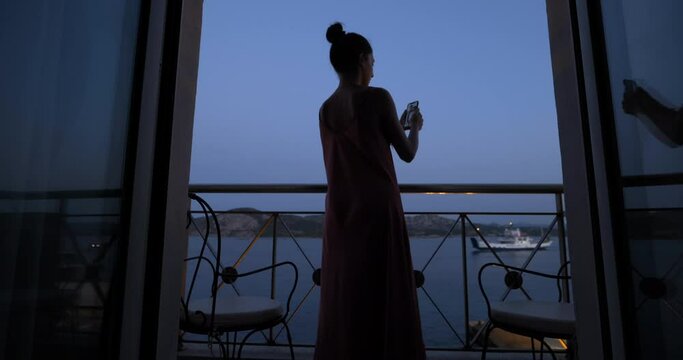 An young woman is taking a picture with smartphone of a panoramic view on a sea with offshore yachts from a terrace of a hotel room during summer holidays vacation trip at sunset