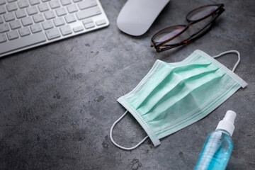 disposable surgical face mask and alcohol spray in working space with computer keyboard, mouse and eyeglasses, concept of flu prevention during situation of COVID-19 to stop pandemic, selective focus