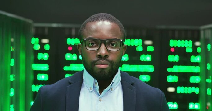 Portrait of African American handsome serious man in glasses looking straight to camera among servers with big data information. Green processors in database storage. Close up of male face of analytic