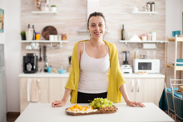 Smiling young woman in kitchen looking at camera with various tasty cheese and grapes on wooden plate.