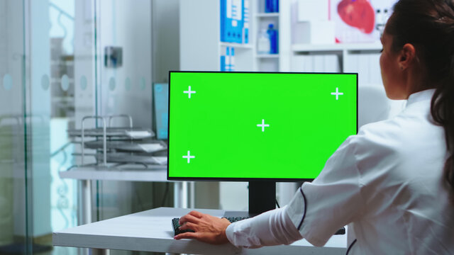 Doctor Sits At Computer With Blank Green Screen Display In Hospital Cabinet And Assistant In Blue Uniform Holding X-ray. Medic In White Coat Working On Monitor With Chroma Key In Clinic Cabinet To