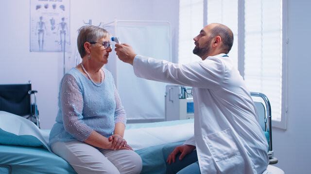 Doctor measuring temperature to senior woman who sits on hospital bed. Using digital infrared contactless thermometer in modern private clinic. Medical examination for infections and disease