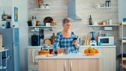 Senior woman browsing on mobile in kitchen during breakfast and smiling to the camera. Authentic elderly person using modern smartphone internet technology. Online communication connected to the world