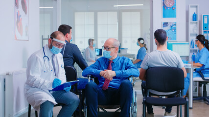Doctor wearing face mask and visor against infection with coronavirus while talking with disabled senior man in waiting area sitting on wheelchair. Patient and nurse at clinic reception.