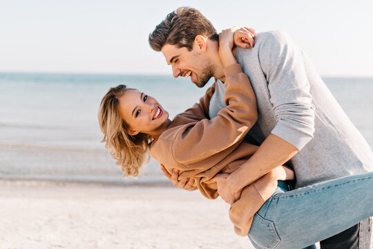 Short-haired blonde lady embracing husband on sea background. Outdoor photo of good-humoured man dancing with girlfriend near ocean.