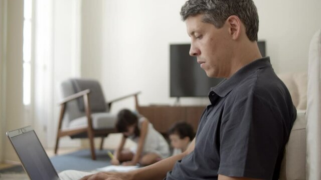 Focused dad with laptop working at home, sitting on floor and using computer while two kids drawing in background. Working at home and parenthood concept