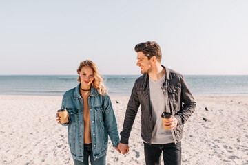 Shy girl holding hands with boyfriend during photoshoot on nature background. Stunning fair-haired lady drinking coffee at sea.