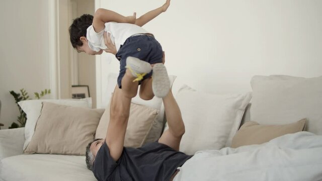 Joyful dad lying on couch, holding and lifting son in air. Boy and his father playing and enjoying leisure time together. Family fun time concept