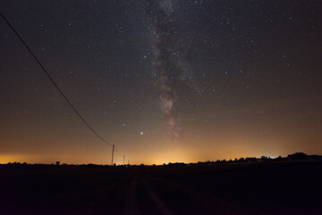 Milky Way on a summer night in Poland. A village is visible on the horizon. Poles with cables break the skyline