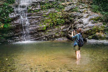 Asian girl tourist traveling adventure exploring nature jungle trail into forest waterfall using camera taking pictures, wearing travel gear walking stick, summer hot healthy outdoor freedom lifestyle