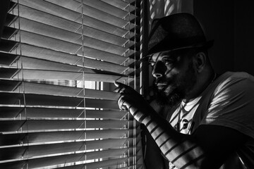 A black man with glasses and a hat looks out the window through the blinds, photographed in black...