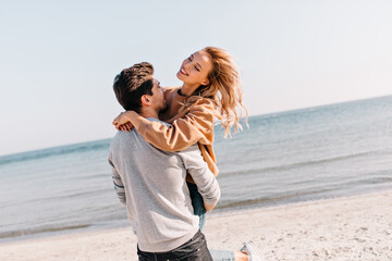 Charming young woman embracing with boyfriend on sea background. Outdoor photo of happy couple standing near ocean.