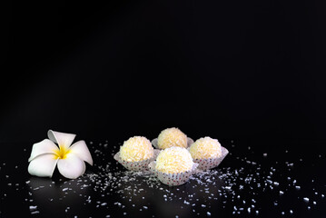 Fototapeta na wymiar coconut flakes scattered around the candy, sweet milk balls with coconut flakes and frangipani flower for decoration, oriental sweets on a black background