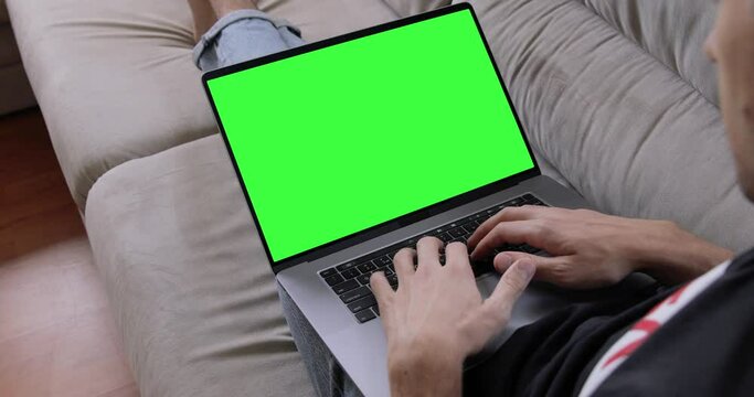 Man sitting on sofa in front of laptop with green screen, chroma key, makes video call to friends, family, working on MacBook Pro keyboard