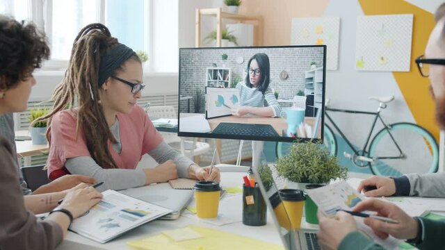 Men and women are talking to female colleague online through video call in office looking and pointing at screen discussing business. People and communication concept.