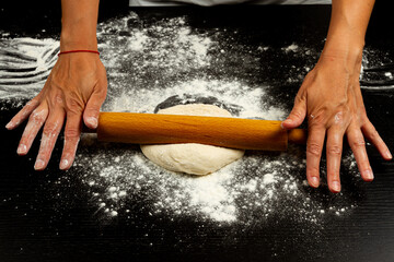 girl cook rolls the dough for homemade pizza with a wooden rolling pin on a black table sprinkled with flour
