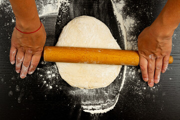 girl cook rolls the dough for homemade pizza with a wooden rolling pin on a black table sprinkled with flour