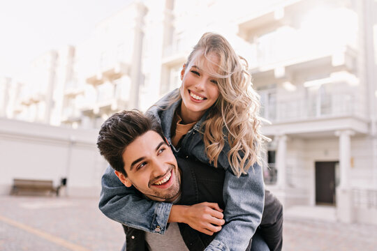 Blithesome fair-haired girl embracing boyfriend with smile. Outdoor photo of funny couple posing on city background.