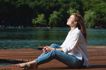 A beautiful and sexy woman, with long hair in a white shirt and jeans, spends time on the shore of the lake, surrounded by forests and mountains. Rest and relaxation concept