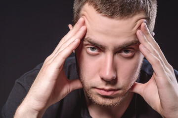 Fototapeta premium Young handsome man in a black shirt sits and holds his head on a black background. Experiencing stress, pain