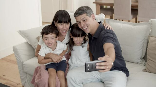 Happy couple of parents with two little kids posing for selfie, sitting on couch in living room, holding phone, smiling, at webcam, waving hands. Technology concept