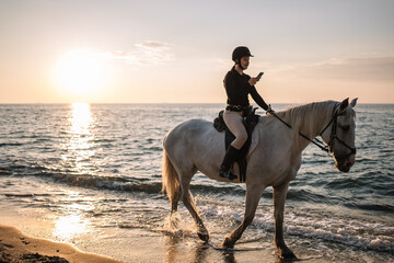 
Woman with phone sending text messages riding on a white horse at sunset