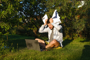 A male manager works remotely at home, makes reports. He is sitting on the lawn in the garden with a laptop. The man is perplexed, holding his head. He is wearing a shirt with a tie, shorts. Stay home