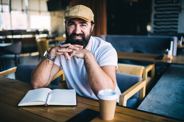 Half length portrait of bearded mature male enjoying spending time in cafe on coffee break smiling businessman looking at camera using notebook with blank pages for writing and planning indoors