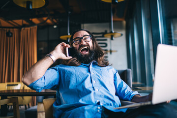 Joyful hipster male with laptop talking on cellphone in cafe