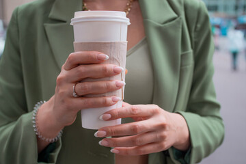 young brunette woman in a green jacket on a walk. Beautiful girl with takeaway coffee with hands