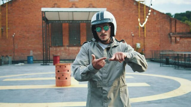 Stylish man in overalls, helmet and sunglasses dancing on the roof gesturing hands looking at camera. Confident talented dancer performing dance elements with modern bricked exterior on background. 4K